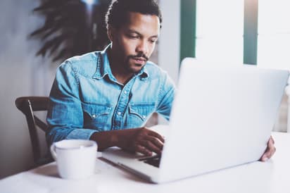 Pensive bearded African-American man working at home while sitting the wooden table.Using modern laptop for new job search.Concept of young people work mobile devices.Blurred background.